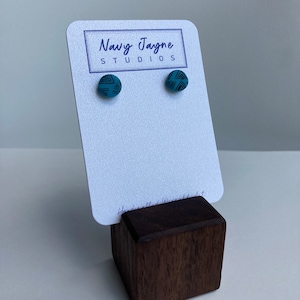 May include: A pair of teal blue stud earrings with a black geometric pattern. The earrings are displayed on a white card with the text "Navy Jayne Studios".