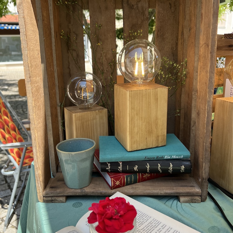 May include: Two wooden lamp bases with clear glass light bulbs, stacked on top of books. The books are on a wooden surface with a blue tablecloth. A red rose is in the foreground.