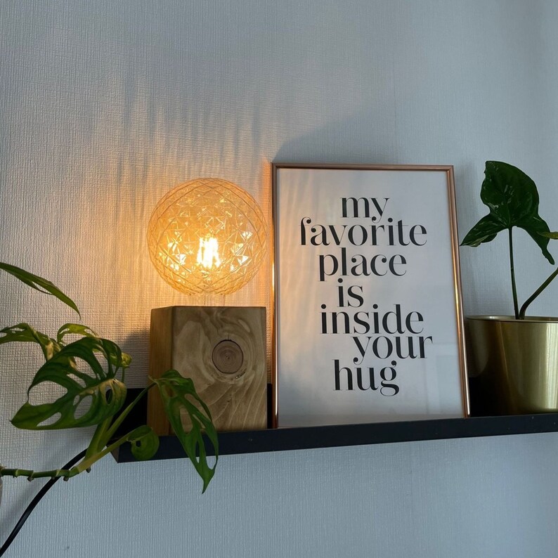 May include: A wooden lamp base with a clear glass globe light bulb. The lamp is on a black shelf with a framed print that reads "my favorite place is inside your hug".