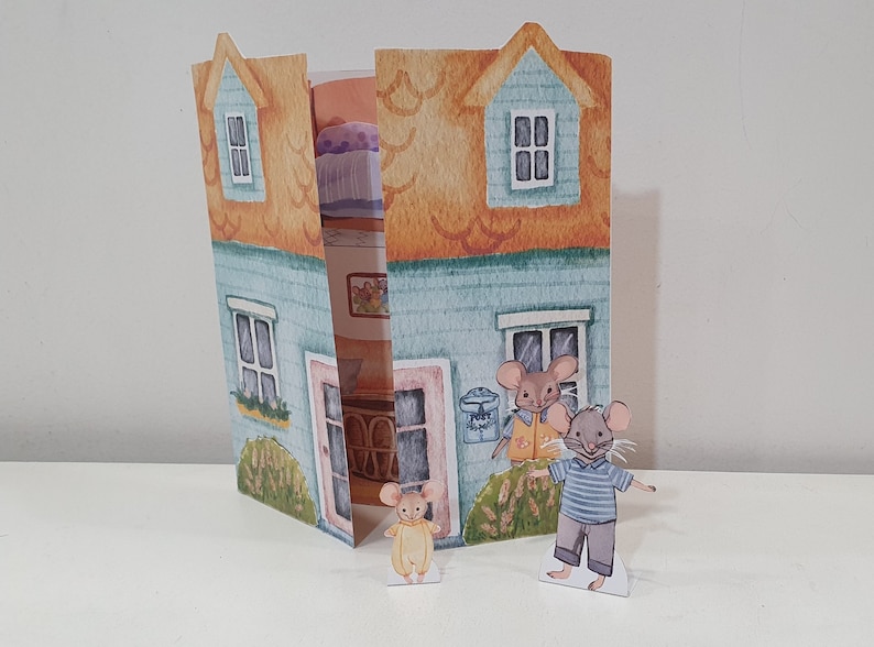 May include: A blue and yellow paper house with a pop-up interior. Two small mice figurines stand in front of the house. One mouse is wearing a yellow shirt and the other is wearing a blue and white striped shirt.