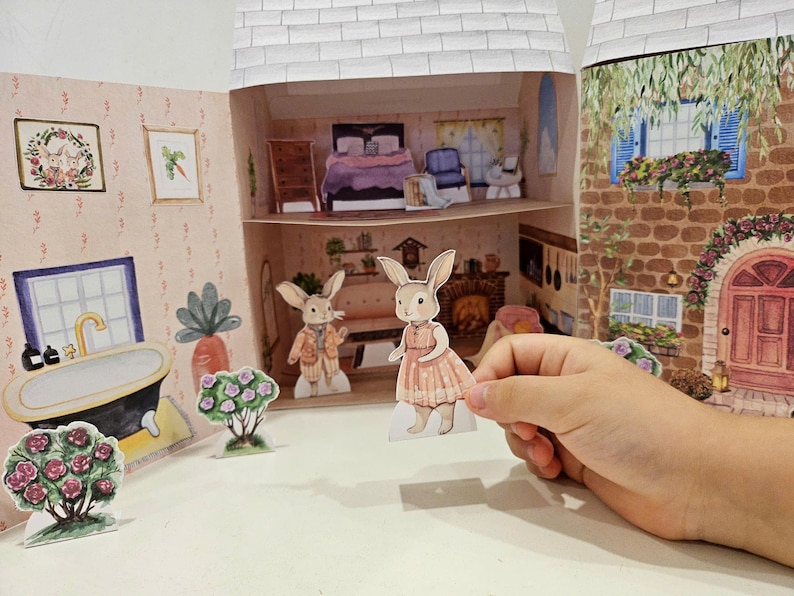 May include: A paper dollhouse playset with two bunny paper dolls, one wearing a pink dress and the other wearing a brown jacket and pants. The dollhouse has a bathroom, bedroom, and kitchen. The dollhouse is made of cardboard and is decorated with colorful illustrations.