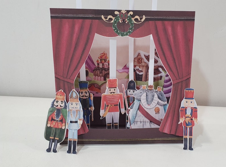 May include: A pop-up card featuring a Nutcracker themed scene with a red curtain, a stage, and several nutcracker figurines. The scene includes a mouse king, a soldier, and a castle.