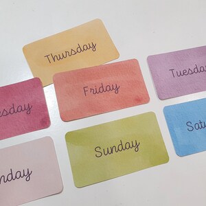Pu&ograve; includere: Sette carte colorate con i giorni della settimana scritti su di esse. Le carte sono disposte a semicerchio su una superficie bianca.