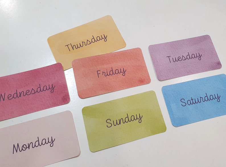 Pu&ograve; includere: Sette carte colorate con i giorni della settimana scritti su di esse. Le carte sono disposte a semicerchio su una superficie bianca.