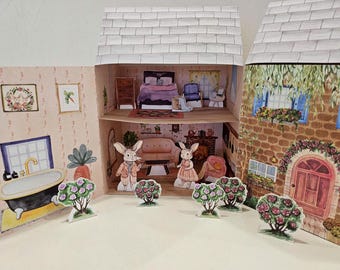May include: A paper dollhouse with two bunny rabbits standing in front of it. The dollhouse has a pink and white exterior with a brown roof. The bunnies are wearing clothes and have pink flowers in front of them.