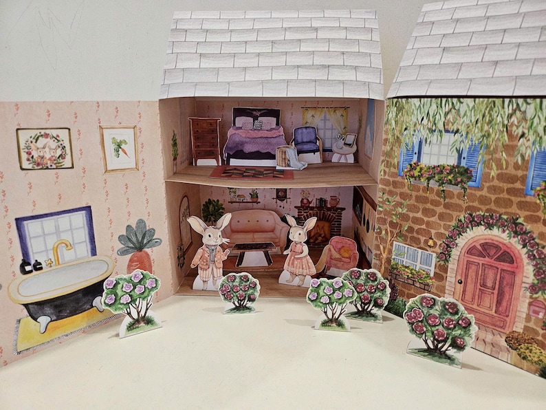 May include: A paper dollhouse with two bunny rabbits standing in front of it. The dollhouse has a pink and white exterior with a brown roof. The bunnies are wearing clothes and have pink flowers in front of them.
