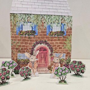 May include: A papercraft model of a house with a red door and two bunnies standing in front of it. The house has a brick facade and two windows with blue shutters. The bunnies are wearing clothes and have flowers around them.