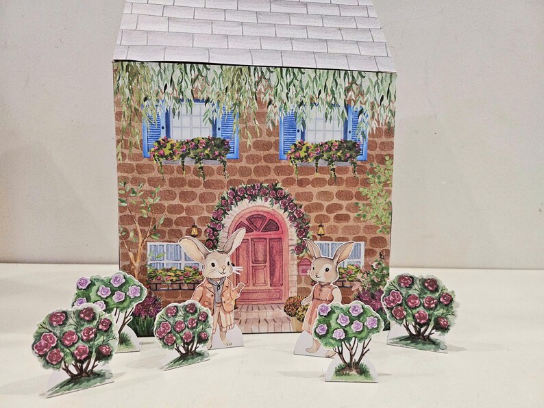 May include: A papercraft model of a house with a red door and two bunnies standing in front of it. The house has a brick facade and two windows with blue shutters. The bunnies are wearing clothes and have flowers around them.
