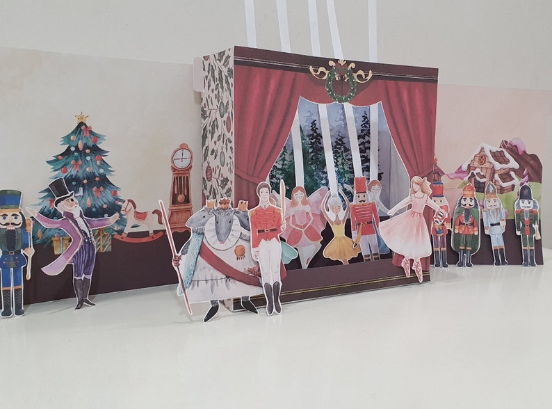 May include: A Nutcracker themed paper craft set with a pop-up stage, a Christmas tree, a clock, a mouse king, a nutcracker soldier, and several dancers. The stage has a red curtain and a window with a snowy forest scene.