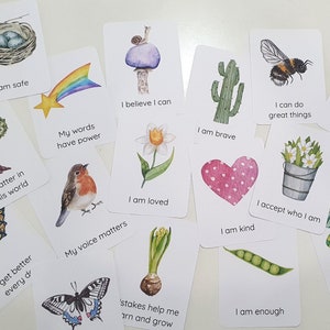 Puede incluir: Un juego de 15 tarjetas de afirmaci&oacute;n con ilustraciones coloridas y mensajes positivos. Las tarjetas presentan im&aacute;genes de un arco&iacute;ris, una oruga, una mariposa, un p&aacute;jaro, un coraz&oacute;n, una flor, un cactus, un hongo, una abeja, un tr&eacute;bol, una vaina de guisante, un caracol, un nido y una planta en maceta. Las tarjetas est&aacute;n impresas con frases como "Estoy seguro", "Soy amado", "Soy valiente", "Puedo hacer grandes cosas", "Importo en este mundo", "Mis palabras tienen poder", "Mi voz importa", "Creo que puedo", "Acepto qui&eacute;n soy", "Soy amable", "Soy suficiente", "Puedo marcar la diferencia", "Me pongo mejor cada d&iacute;a", "Estoy agradecido por hoy", y "Las estacas me ayudan a aprender y crecer".