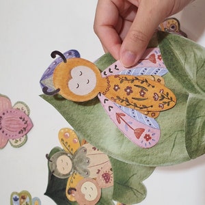 May include: A set of colorful paper cutouts of butterflies and a leaf. The butterflies have floral patterns and are in shades of pink, purple, yellow, and green. The leaf is green and has a watercolor effect.