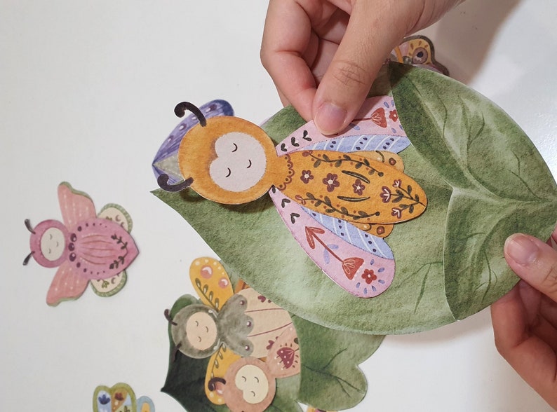 May include: A set of colorful paper cutouts of butterflies and a leaf. The butterflies have floral patterns and are in shades of pink, purple, yellow, and green. The leaf is green and has a watercolor effect.