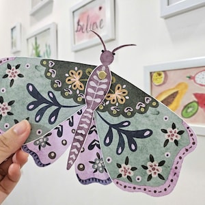 Puede incluir: Una mariposa de papel con un diseño verde y morado. La mariposa tiene un patrón floral y está decorada con detalles dorados.