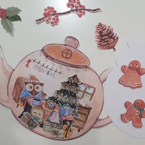 以下が含まれることがあります： ピンクのティーポットに茶色の蓋と白い取っ手。ティーポットはクリスマスツリー、窓、暖炉で飾られています。また、フクロウ2羽、ジンジャーマン、松ぼっくりも描かれています。