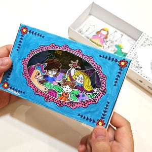 Puede incluir: Una caja de arte de papel hecha a mano con una escena de cuento de hadas. La caja tiene un fondo azul con un marco ovalado rosa, y la escena incluye un unicornio, hadas y una princesa. Se ven recortes de papel adicionales en la caja abierta.