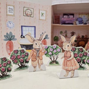 May include: Two paper cutouts of bunnies dressed in clothes stand in front of a miniature dollhouse. The bunny on the left is wearing a brown and orange jacket and pants. The bunny on the right is wearing a pink dress. Both bunnies are standing in front of bushes with pink flowers.