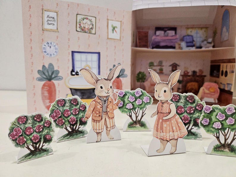 May include: Two paper cutouts of bunnies dressed in clothes stand in front of a miniature dollhouse. The bunny on the left is wearing a brown and orange jacket and pants. The bunny on the right is wearing a pink dress. Both bunnies are standing in front of bushes with pink flowers.