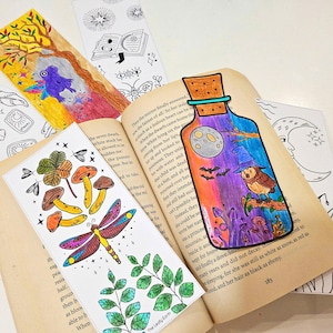 May include: A collection of colorful, hand-drawn bookmarks. One features a potion bottle with an owl, moon, and bats. Others depict a tree with a bird, a dragonfly with mushrooms, and celestial designs. The bookmarks are placed in an open book.