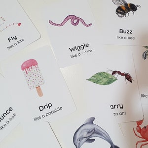 May include: A collection of white flashcards with watercolor illustrations and text. The cards feature images of a kite, worm, bee, popsicle, ball, ant, dolphin, palm tree, and crab. Each card has a word and a phrase like "Fly like a kite."