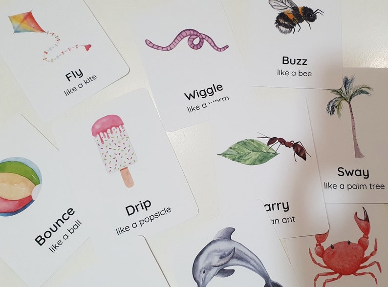 May include: A collection of white flashcards with watercolor illustrations and text. The cards feature images of a kite, worm, bee, popsicle, ball, ant, dolphin, palm tree, and crab. Each card has a word and a phrase like "Fly like a kite."