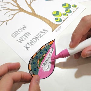 May include: A person coloring a leaf-shaped paper cutout with a pink marker. The cutout has a decorative design and the text "That was nice of you". The words "Grow with Kindness" are printed on the paper.