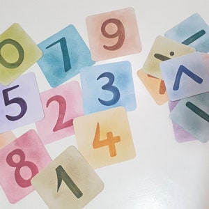 May include: A set of colorful, square cards with numbers 1 through 9 and math symbols, including plus, minus, and division signs. The cards are made of thick paper and have a watercolor-like finish.