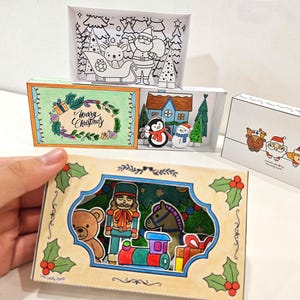 May include: A collection of handmade Christmas-themed paper art boxes. The boxes feature colorful illustrations of Santa, reindeer, snowmen, and other festive characters. One box is held by a hand, showcasing a nutcracker and toy train.