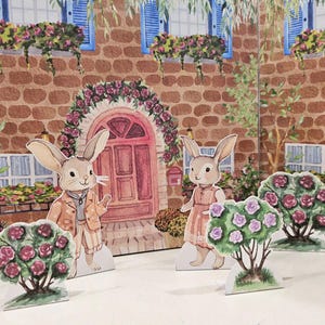 May include: Two paper cutouts of rabbits dressed in clothing stand in front of a brick building with windows and a red door. The building is decorated with flowers and vines. The rabbits are surrounded by bushes with pink flowers.