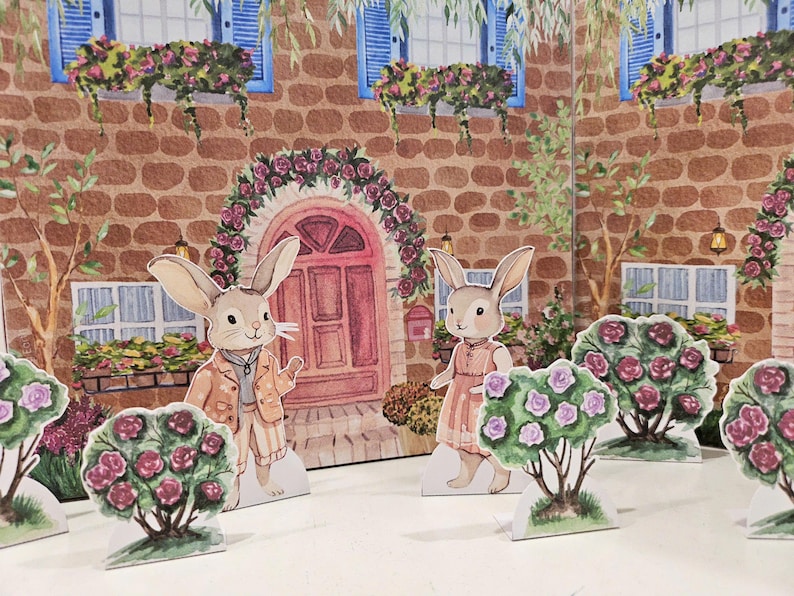 May include: Two paper cutouts of rabbits dressed in clothing stand in front of a brick building with windows and a red door. The building is decorated with flowers and vines. The rabbits are surrounded by bushes with pink flowers.