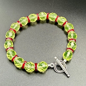 May include: A beaded bracelet featuring faceted, light green beads separated by red rhinestone accents. The bracelet has a silver-tone toggle clasp. The beads are round and have a sparkling, crystal-like appearance.