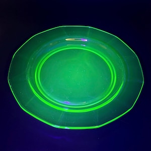 May include: A glowing green glass plate with a unique octagonal shape. The plate has a smooth, reflective surface and a bright, neon-like appearance. The plate is set against a dark blue background.