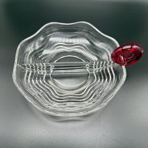 Könnte beinhalten: Eine klare Glasservierschale mit wellenförmigem Design und rotem Griff. Die Schale ist in zwei Abschnitte unterteilt und eignet sich zum Servieren von Snacks oder Vorspeisen. Der Griff ist tiefrot.