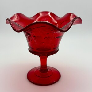 Op de afbeelding: Een robijnrode glazen voetkom met een gegolfde rand. De kom heeft een gedetailleerd bloemmotief en staat op een ronde voet die door een steel is verbonden. Deze vintage kom is geschikt voor het serveren van desserts of het tentoonstellen van decoratieve items.