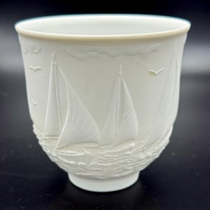 Könnte beinhalten: Weiße Keramiktasse mit nautischem Design. Die Tasse zeigt geprägte Segelboote auf einem welligen Meer, mit Möwen und Wolken. Der Rand hat eine hellbeige Farbe.