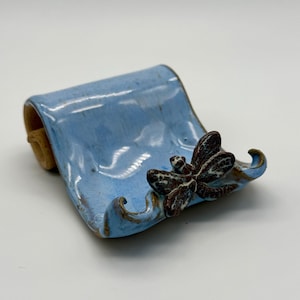 May include: A ceramic soap dish with a light blue glaze. It features a sculpted dragonfly with brown and white details. The edges have a brown trim, and the dish has a curved shape. Made by hand.