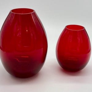 Può includere: Due vasi di vetro rosso, uno più grande dell'altro, con una forma arrotondata a forma di lacrima.