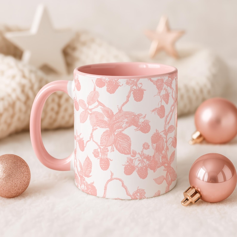 May include: A white ceramic mug with a pink interior and handle. The mug has a pink raspberry design. Pink Christmas baubles and stars are in the background, creating a festive holiday theme.
