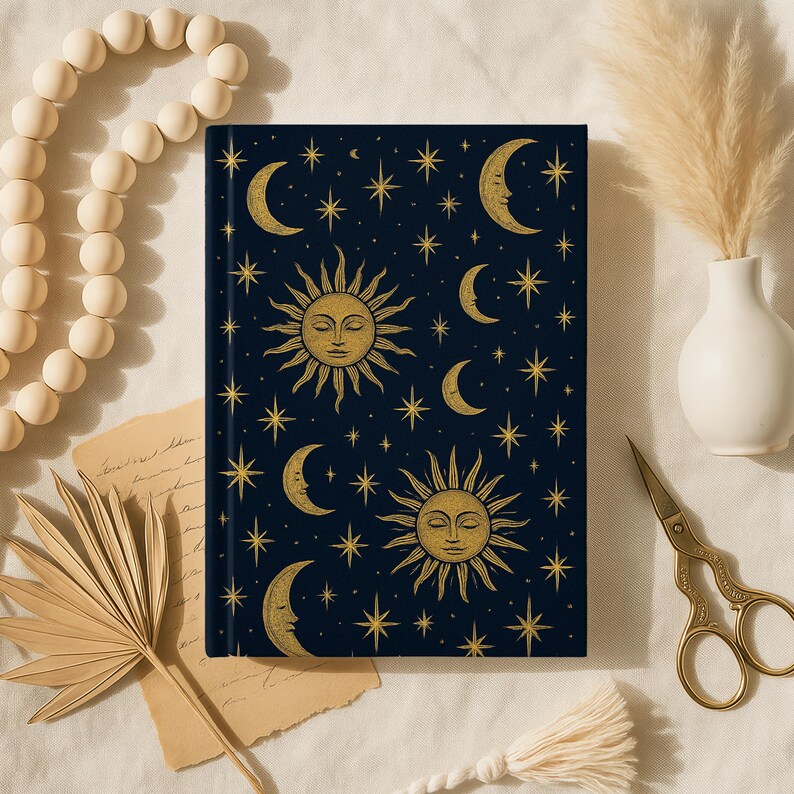 May include: A dark blue journal with a celestial design featuring gold suns, crescent moons, and stars. The journal is surrounded by decorative elements, including a beaded necklace, dried plants, and gold scissors.