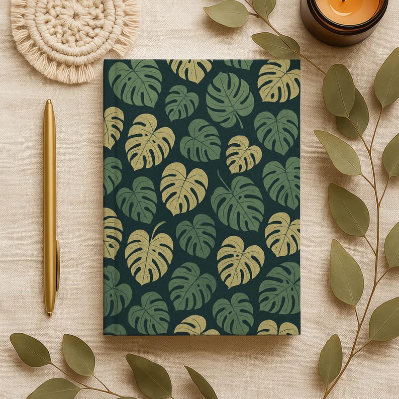 May include: A notebook with a dark teal background and a pattern of green and light green monstera leaves. A gold pen and a beige coaster are on the left. A lit candle and greenery are on the right.