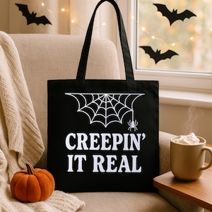 Op de afbeelding: Zwarte draagtas met een wit spinnenweb en spinnenafbeelding, en de tekst "CREEPIN' IT REAL". Een oranje pompoen en een mok slagroom zijn ook zichtbaar. De tas heeft zwarte handvatten en staat op een beige stoel.