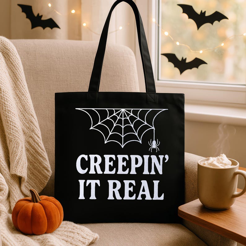 Op de afbeelding: Zwarte draagtas met een wit spinnenweb en spinnenafbeelding, en de tekst "CREEPIN' IT REAL". Een oranje pompoen en een mok slagroom zijn ook zichtbaar. De tas heeft zwarte handvatten en staat op een beige stoel.
