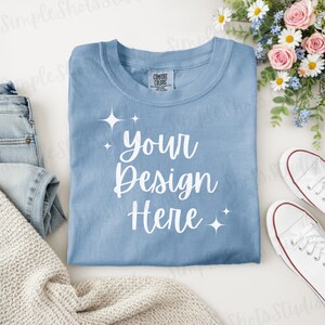Könnte beinhalten: Hellblaues T-Shirt mit dem weißen Schriftzug "Your Design Here". Das Shirt ist gefaltet und von einer Jeans, einem cremefarbenen Pullover, weißen Turnschuhen und Blumen umgeben.