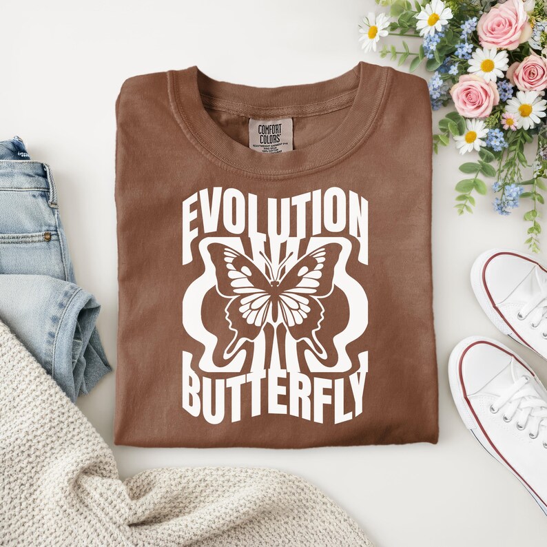 Puede incluir: Una camiseta marr&oacute;n con la inscripci&oacute;n blanca "EVOLUTION BUTTERFLY" y un dise&ntilde;o de mariposa. La camiseta est&aacute; doblada sobre una superficie blanca, con zapatillas blancas y un ramo de flores en el fondo.