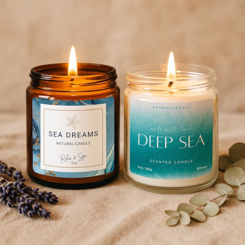 May include: Two lit scented candles in glass jars. One is amber-colored with a label reading "Sea Dreams" and the other is clear with a label reading "Deep Sea." Both labels have blue and white designs. The "Deep Sea" candle is 8 oz/300g.