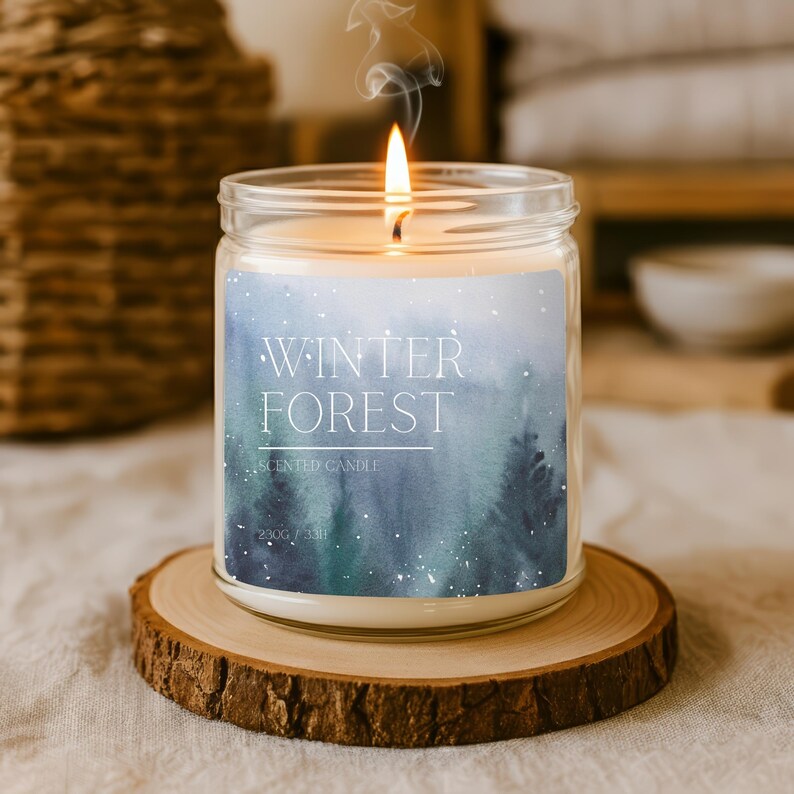 Op de afbeelding: Een brandende geurkaars in een glazen pot met een etiket met de tekst "Winter Forest". Het etiket toont een aquarelontwerp van een bos in blauw- en groentinten. De kaars staat op een houten onderzetter. De kaars weegt 230g.
