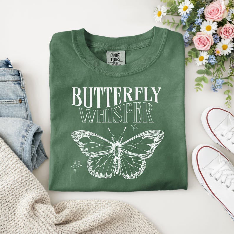 Puede incluir: Camiseta verde con la frase "BUTTERFLY WHISPER" y un dibujo de mariposa blanca. La camiseta est&aacute; doblada, con unos vaqueros azules y un su&eacute;ter color crema a la izquierda. Zapatillas blancas y flores a la derecha.