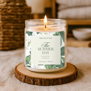May include: A lit scented candle in a glass jar with a label that reads "The Summer Day." The label has a floral design with green leaves and white flowers. The candle sits on a wooden slice and is labeled "40 hours / 300 g."