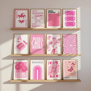 May include: A collection of framed pink and white art prints displayed on wooden shelves. The prints feature various designs, including playing cards, dice, text, and abstract patterns. The text includes "JOKER", "More Espresso", "Perfect Match", "Chill", "Feeling Lucky", "BAUHAUS", "GOOD TIMES", "YAYOI KUSAMA", and "KAMISAKA SEKKA".