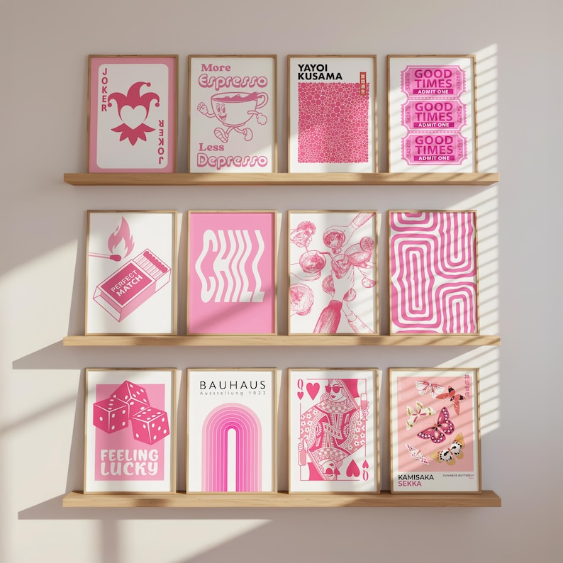 May include: A collection of framed pink and white art prints displayed on wooden shelves. The prints feature various designs, including playing cards, dice, text, and abstract patterns. The text includes "JOKER", "More Espresso", "Perfect Match", "Chill", "Feeling Lucky", "BAUHAUS", "GOOD TIMES", "YAYOI KUSAMA", and "KAMISAKA SEKKA".