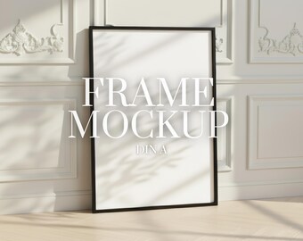 May include: A black framed art print mockup leaning against a white wall with decorative molding. The text "FRAME MOCKUP DINA" is displayed on the white print. The floor is light-colored wood.